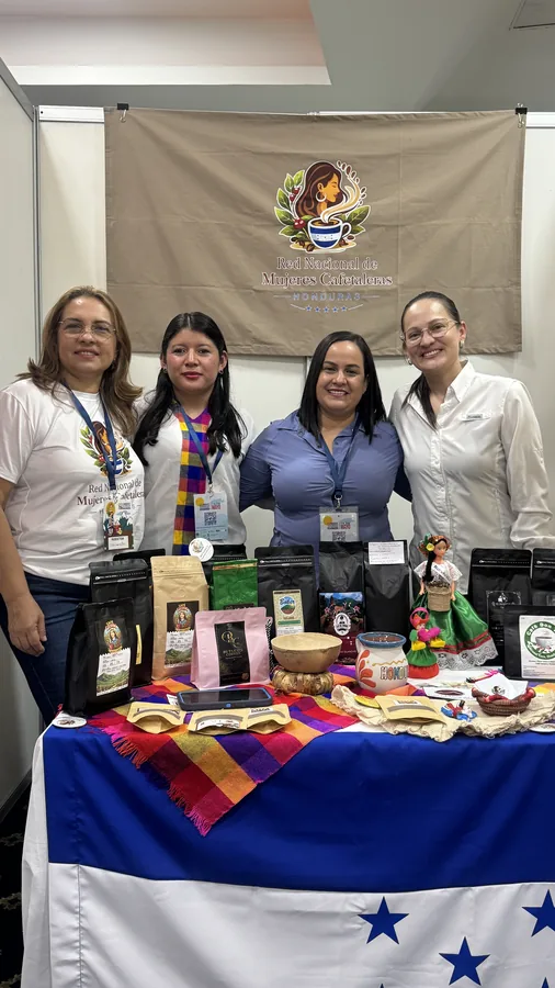 Cuatro mujeres posando en un stand de expo cafetera con bandera de Honduras y productos artesanales