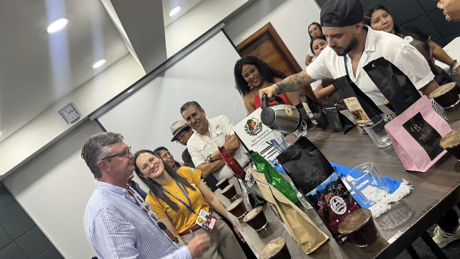 Mesa amplia de catación con tazas y bolsas de café, personas sentadas y de pie alrededor