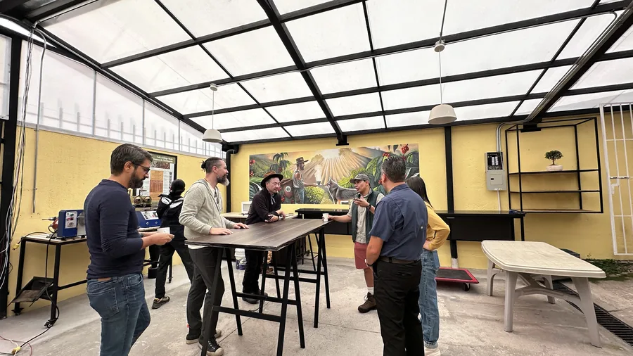 Grupo conversando dentro del laboratorio de café bajo techo de vidrio, con el mural de la finca al fondo