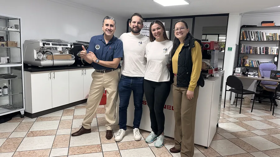 Cuatro colegas posando juntos en la oficina de equipos, con una máquina de espresso y una biblioteca de fondo