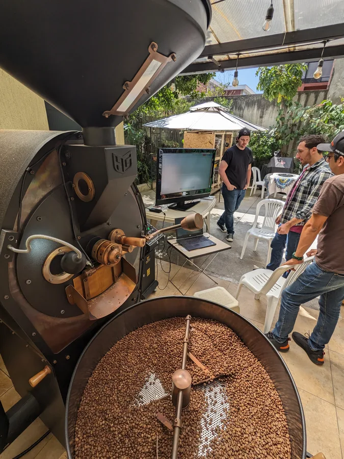 Tostadora en primer plano con el monitor de curva de tueste en vivo detrás, personal observando bajo luces de patio cubierto
